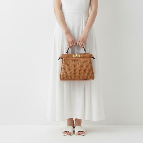 Model holding brown woven vegan leather satchel bag with gold clasp, wearing a white dress