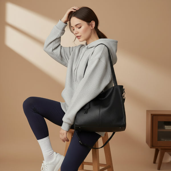 Side view of a woman wearing a gray hoodie and black leggings, holding a stylish black vegan leather tote bag with a decorative tassel
