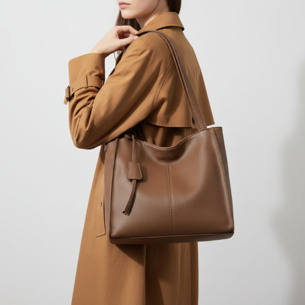 Side view of a woman wearing a tan coat, holding a brown vegan leather tote bag with a sleek design and a decorative leather tassel