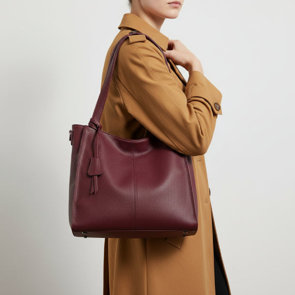 Side view of a woman wearing a beige trench coat, holding a stylish wine red vegan leather tote bag with a sleek design and decorative tasse