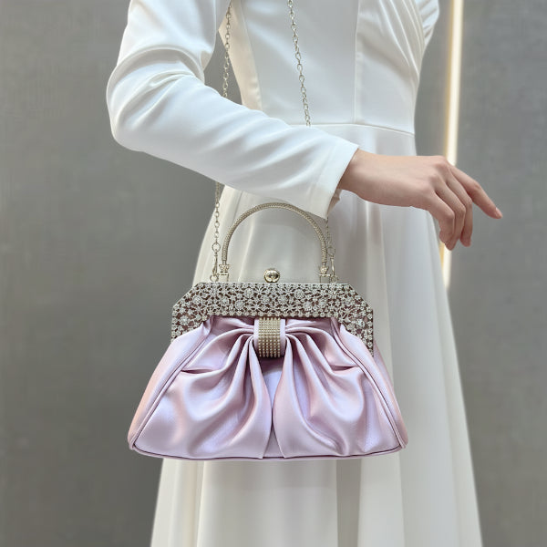 Model holding a pink vegan leather evening bag with rhinestone detailing, pleated satin fabric, and a gold frame, styled with a white dress