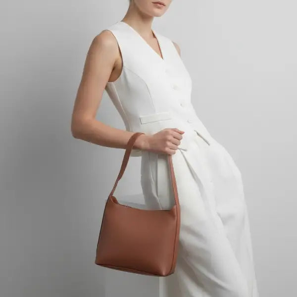 Model wearing a white outfit, holding a brown vegan leather shoulder bag with a gold buckle on the strap