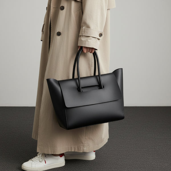 Model carrying black vegan leather tote bag with flap closure, styled with a beige trench coat and white sneakers for a chic, casual appearance