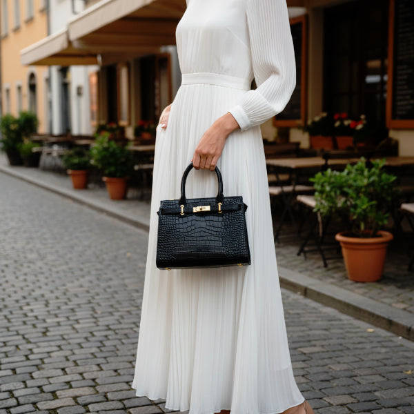 Model holding Adara genuine leather black satchel bag while wearing a white dress in an outdoor setting