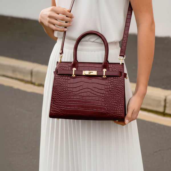 Model holding Adara genuine leather wine red satchel bag with gold hardware, styled with a white dress in an outdoor setting