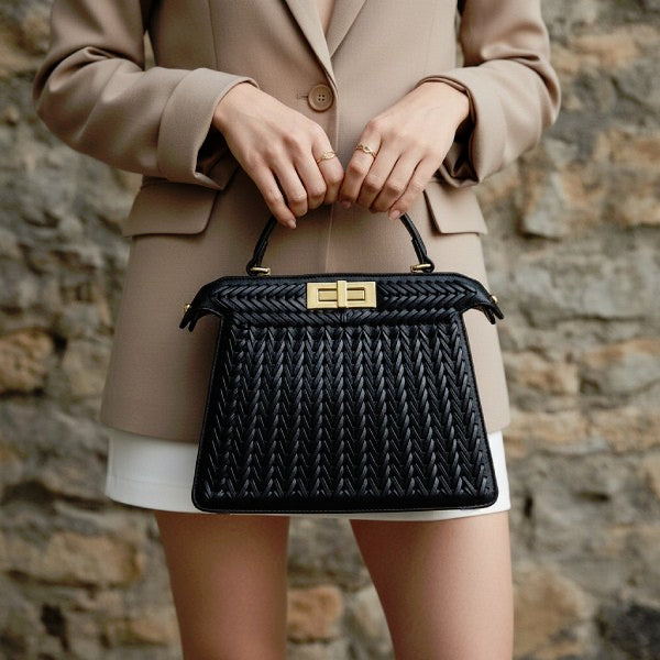 Model holding black woven vegan leather satchel bag with gold clasp and handle, wearing beige blazer