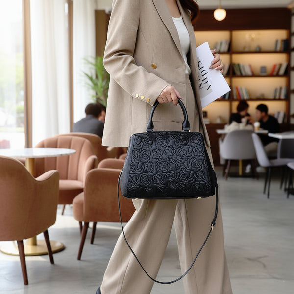 Model holding Azura black leather satchel bag while walking inside a café, paired with a beige suit