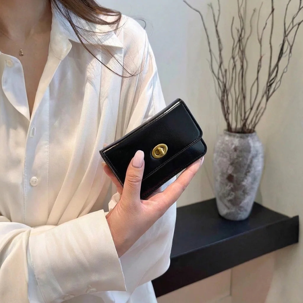 Person holding a black wallet with a gold clasp in a minimalistic indoor setting.