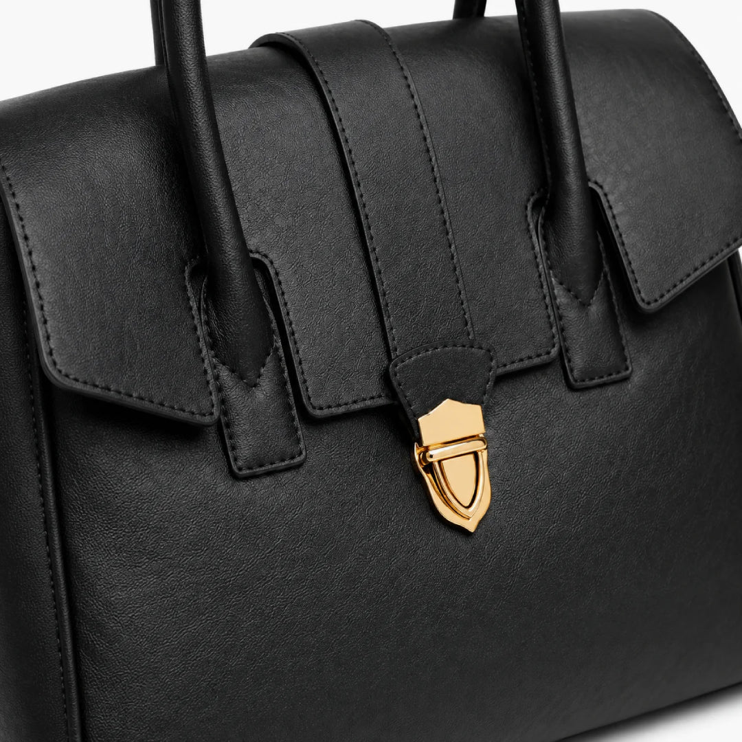 Close-up of a black textured leather handbag with a gold shield-shaped clasp and black handles on a white background.