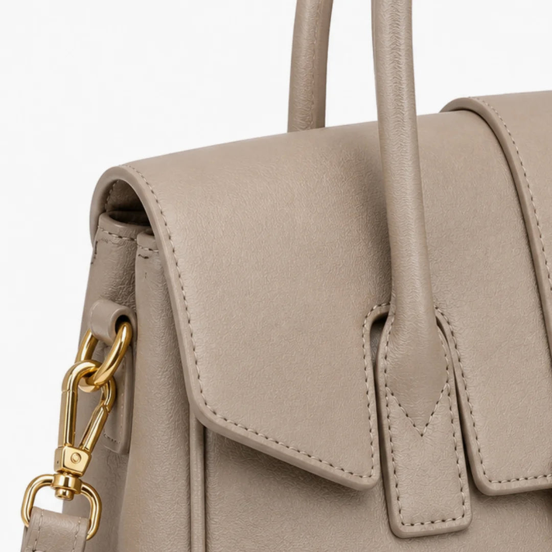 Close-up of a taupe leather handbag with gold-tone hardware, showing the flap, handles, and clasp against a white background.