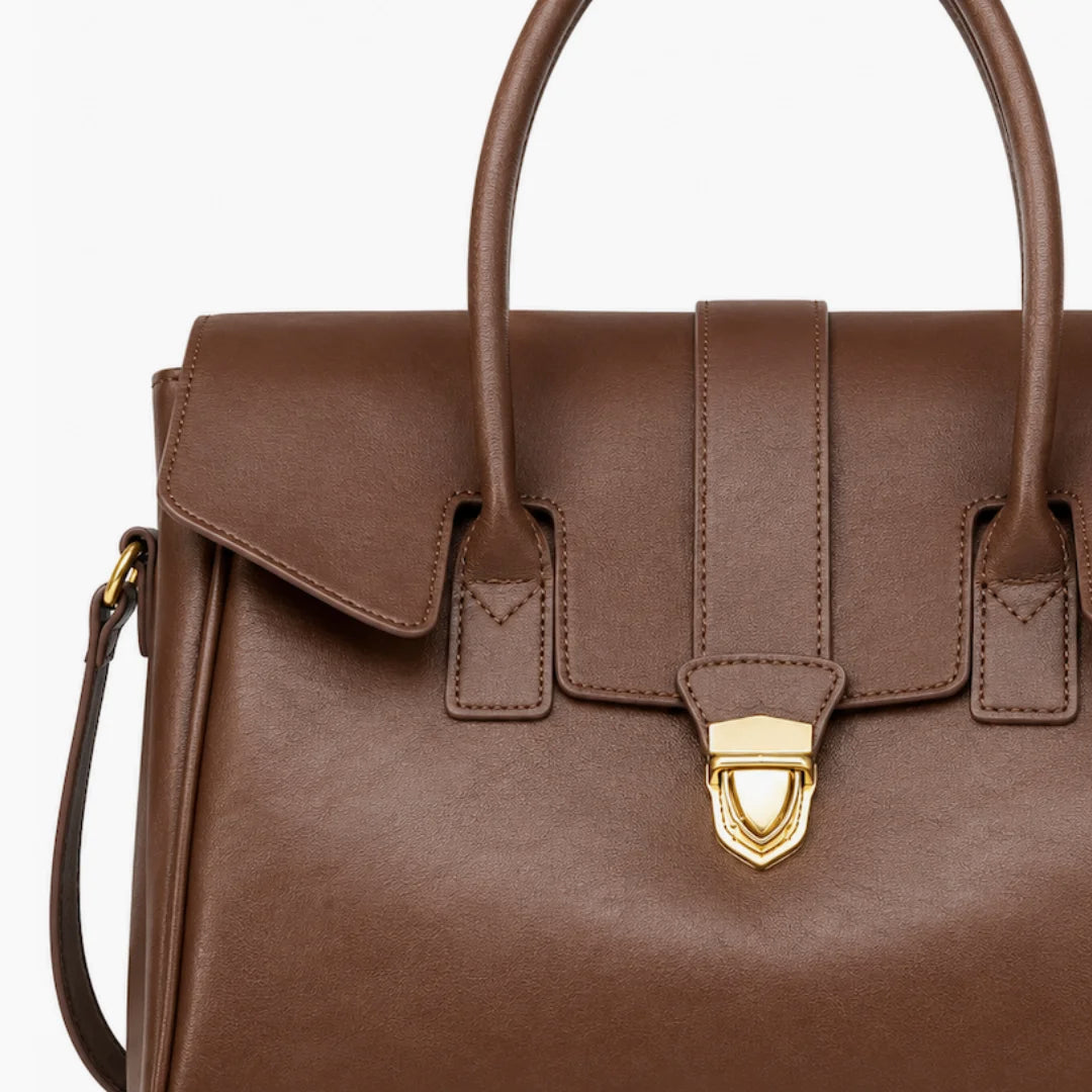 A close-up of a brown leather handbag featuring a structured top handle, a front flap with a central strap, and a gold-tone shield-shaped clasp on a white background.
