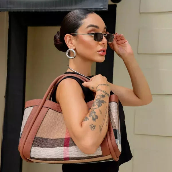 Woman with tattoos holding a checkered handbag, wearing sunglasses, in front of a house.