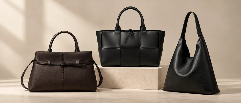 Three handbags on a beige stone pedestal: a dark brown satchel, a black woven tote and a black hobo bag