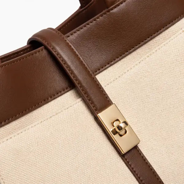 Close-up of a tote bag featuring a cream canvas body, brown leather trim, and a gold-tone metal clasp on a strap.