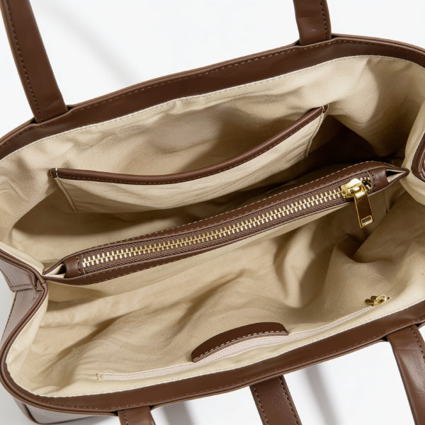 Close-up of the interior of a brown handbag, showing a beige fabric lining, a zippered pocket with a gold-tone zipper pull, and the edge of the bag against a white background.