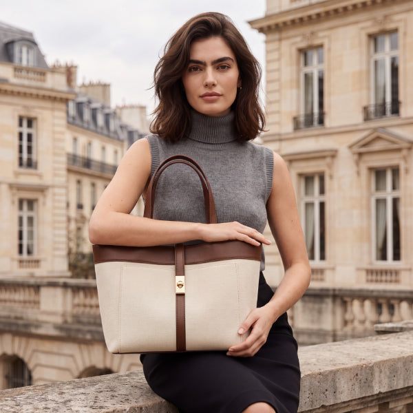 A woman in a gray sleeveless turtleneck sweater and black skirt sits on a stone ledge in Paris, holding a cream and brown leather tote bag with a gold-tone front clasp.