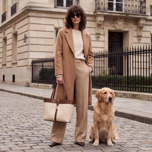 A woman stands on a cobblestone street next to a golden retriever, wearing a camel coat, beige trousers, and holding a cream tote bag with brown leather handles. A grand stone building and black iron fence are in the background.