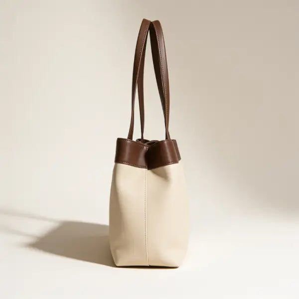 A minimalist leather tote bag with a cream-colored body and dark brown handles and trim, standing on a light beige background.