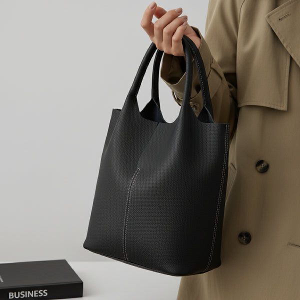 Model holding black cowhide leather tote bag with minimalist design, styled with a beige trench coat for a sophisticated, casual appearance