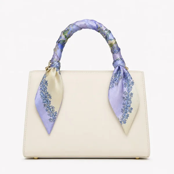 An off-white structured handbag with a decorative silk scarf handle in lavender, blue, and cream tones, set against a plain white background.