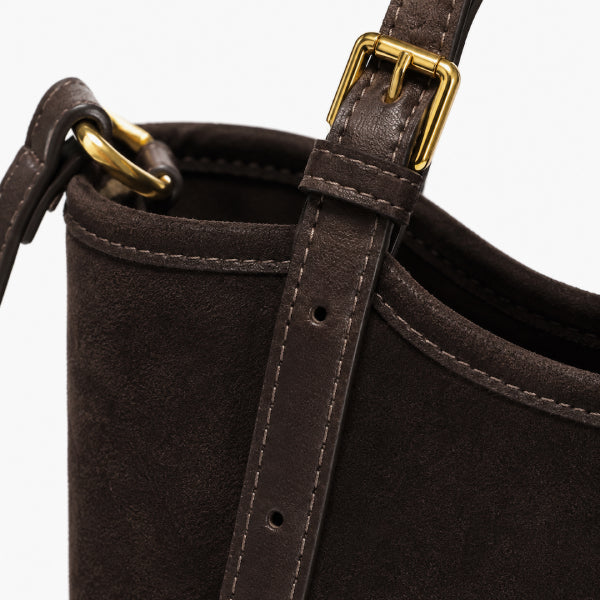 Close-up of a dark brown suede bucket bag with a matching leather strap and gold-tone buckle hardware, shown against a plain light background.
