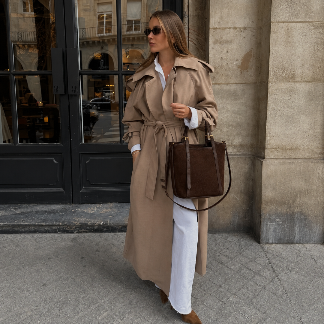 A woman wearing a long beige trench coat over a white shirt and white wide-leg pants, carrying a dark brown leather tote bag, walking on a city sidewalk in front of a stone building.