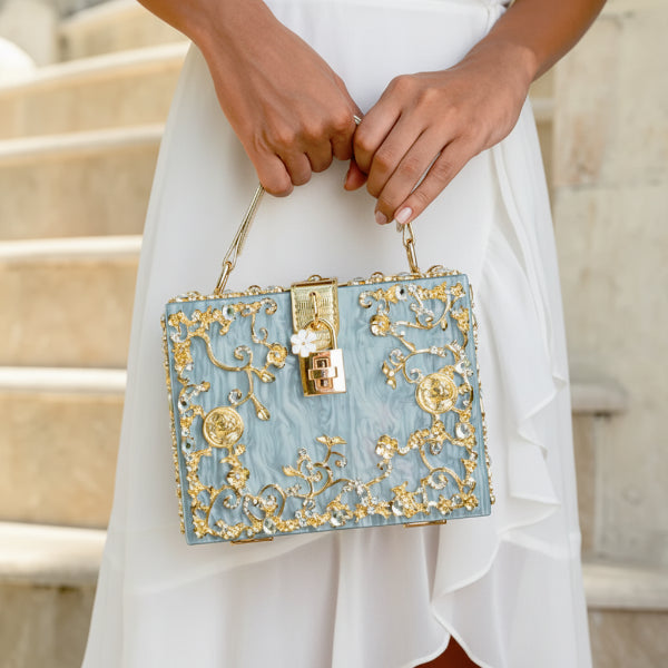 Model holding Zephyra acrylic evening bag in blue with gold floral accents and lock closure